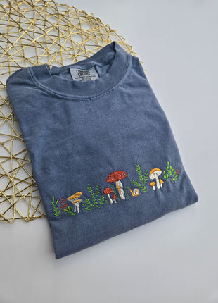 A folded slate blue t-shirt with a vertical embroidered line of colorful mushrooms and green leaves, displayed on a gold wire geometric mat against a light gray background.
