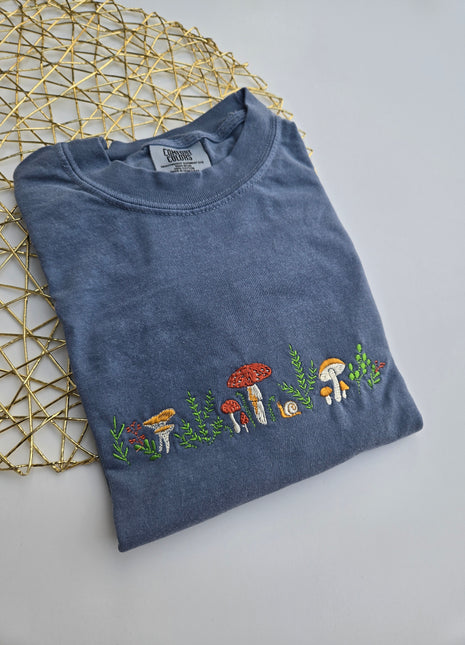 A folded slate blue t-shirt with a vertical embroidered line of colorful mushrooms and green leaves, displayed on a gold wire geometric mat against a light gray background.