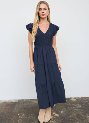 A woman modeling a navy blue midi dress with ruffled cap sleeves, a smocked bodice, and a tiered skirt, standing against a white wall.