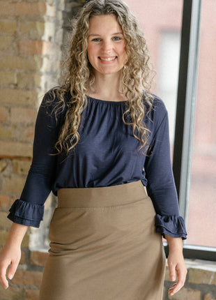 Woman wearing a navy blouse and brown skirt standing indoors with a brick wall background