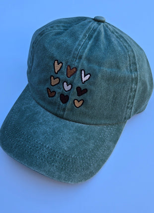 A washed teal-green baseball cap featuring embroidered hearts in various colors on the front, set against a solid light blue background.