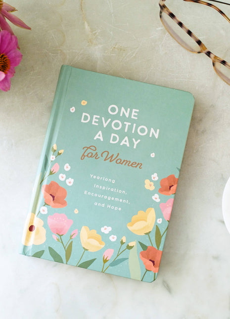 Book titled 'One Devotion A Day for Women' with floral design on a marble surface with flowers and glasses.
