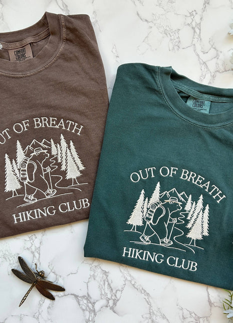 Two t-shirts with 'Out of Breath Hiking Club' design on a marble surface with flowers.