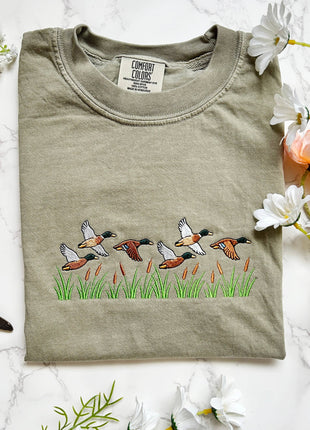 A folded sage green t-shirt with an embroidered duck family scene on the front, displayed on a white marble background with decorative flowers and a dragonfly.