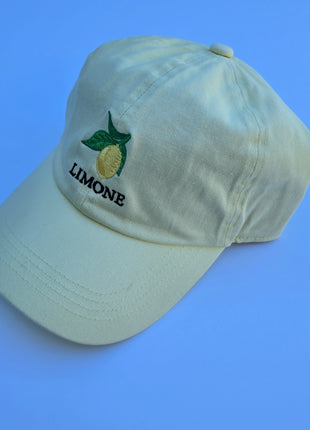 A light yellow cotton baseball cap with an embroidered lemon and the word 'LIMONE' on the front, set against a solid blue background.