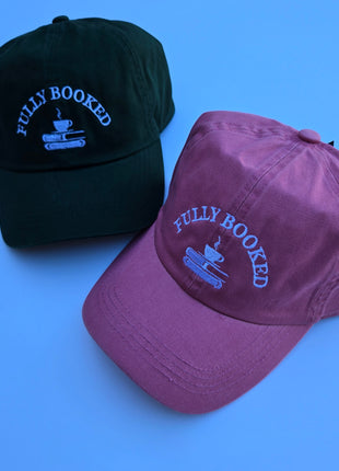 Two embroidered baseball caps on a solid blue background. One cap is dark green with light blue embroidery reading 'FULLY BOOKED' and a coffee cup on books graphic.