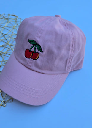 A light purple baseball cap featuring an embroidered cherry design on the front, styled on a blue background next to a gold wire mesh tray.