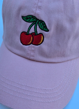 A light pink baseball cap featuring an embroidered design of two red cherries with green leaves on the front panel, set against a solid light blue background.