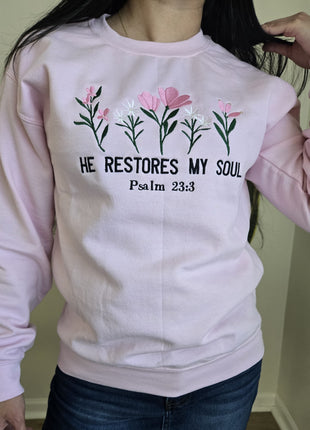 A person wearing a light pink crewneck sweatshirt with a graphic of pink and white flowers, green stems, and the text 'HE RESTORES MY SOUL Psalm 23:3' in black. The person is also wearing dark blue jeans, standing in an indoor setting.