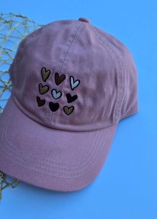 A dusty purple baseball cap with embroidered hearts in various colors on the front, styled on a blue background next to a gold wire mesh prop.