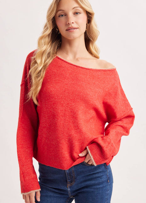 A woman wearing a bright coral-red off-the-shoulder knit sweater and dark blue jeans, photographed against a clean light gray studio background.