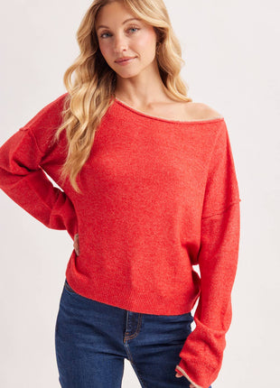 A woman wearing a bright red off-the-shoulder knit sweater and dark blue jeans, photographed against a clean light gray studio background.