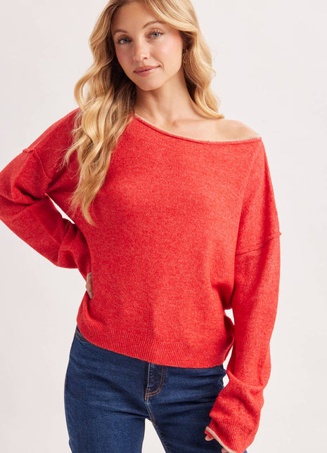 A woman wearing a bright red off-the-shoulder knit sweater and dark blue jeans, photographed against a clean light gray studio background.