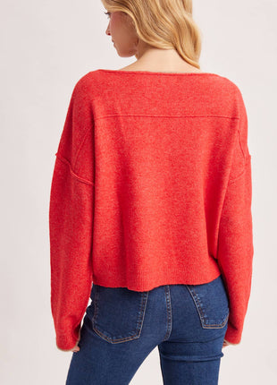 A woman shown from the back wearing a bright coral-red knit sweater and blue jeans against a plain light gray studio background.