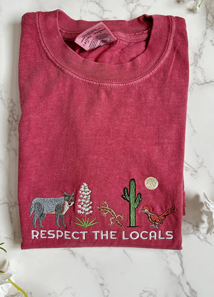 Folded red t-shirt with nature-themed graphics and 'Respect the Locals' text on a marble surface.