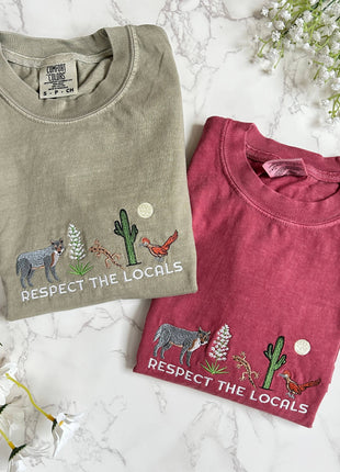 Two folded t-shirts with nature-themed designs and text on a marble surface with flowers.