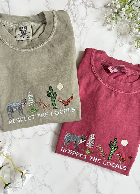 Two folded t-shirts with nature-themed designs and text on a marble surface with flowers.