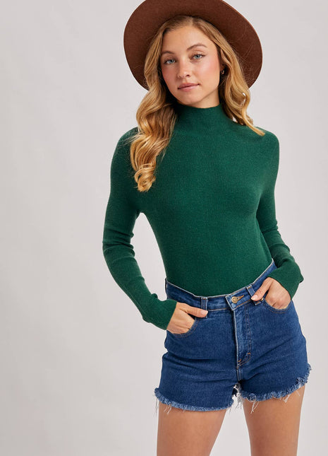 A woman modeling a dark green mock neck knit sweater, paired with blue denim shorts and a brown wide-brim hat, against a light gray studio background.