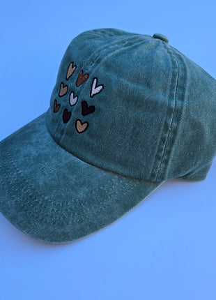 A washed teal baseball cap with embroidered hearts in various colors on the front, set against a solid blue background.