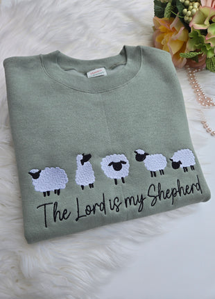 A folded sage green crewneck sweatshirt featuring embroidered white sheep with black faces and the text 'The Lord is my Shepherd' in black script, displayed on a white faux fur surface with decorative flowers and a pearl strand.