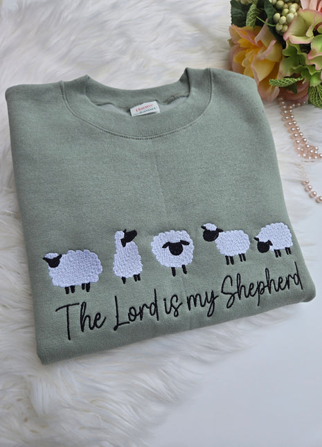 A folded sage green crewneck sweatshirt featuring embroidered white sheep with black faces and the text 'The Lord is my Shepherd' in black script, displayed on a white faux fur surface with decorative flowers and a pearl strand.