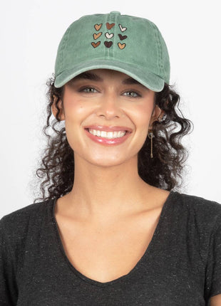 A smiling woman with curly dark hair wearing a green baseball cap with embroidered hearts on the front, paired with a dark gray V-neck top against a plain white background.