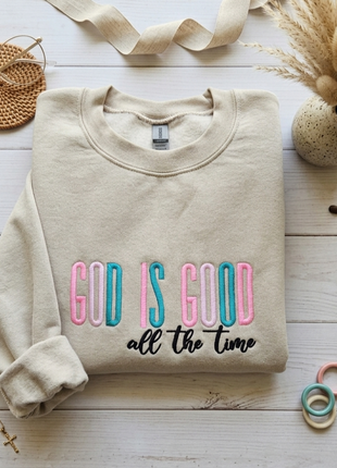 A folded beige crewneck sweatshirt with the phrase 'GOD IS GOOD all the time' printed on the front, styled in a flat lay on a white wood background with decorative props including a woven round mat, dried grass in a pot, and small rings.