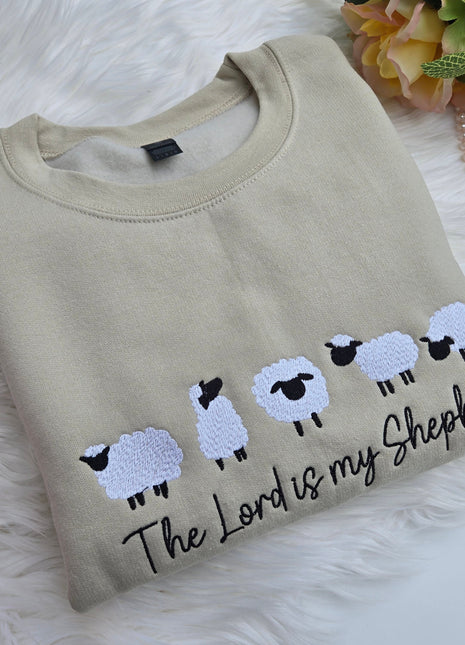 A folded beige crewneck sweatshirt featuring embroidered white sheep with black faces and the phrase 'The Lord is my Shepherd' in black script 