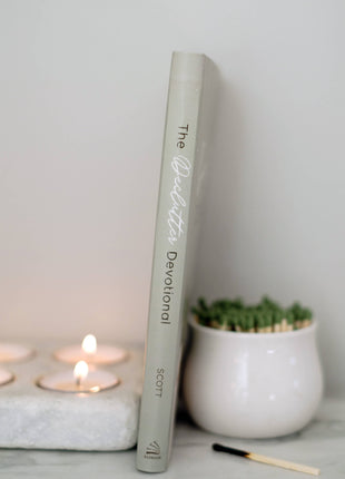 Book titled 'The Devotional' on a surface with candles and a plant in the background