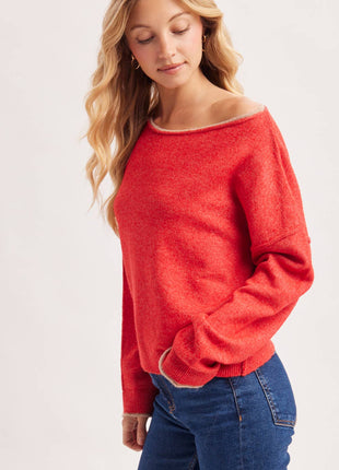 A woman wearing a bright coral-red off-the-shoulder knit sweater and blue jeans, photographed against a clean light gray studio background.