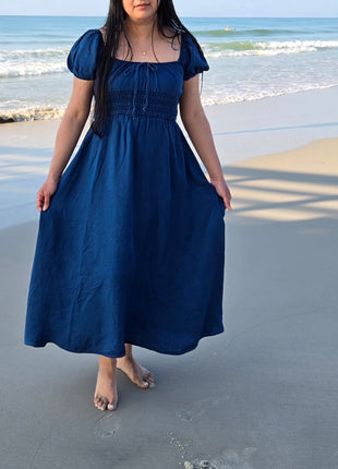 A woman wearing a navy blue midi dress with puff sleeves and a smocked bodice, standing barefoot on a sandy beach with the ocean waves in the background.
