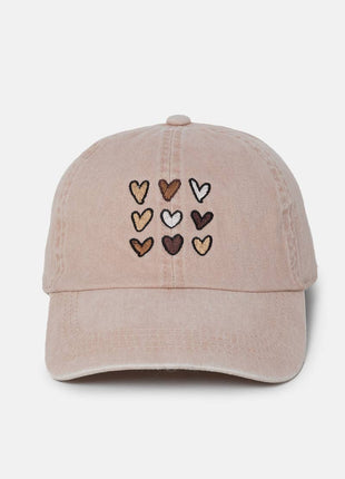 A light pink washed cotton baseball cap featuring nine embroidered hearts in various shades of brown, beige, and white on the front panel, set against a plain light gray background.