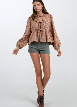 A woman modeling a brown and beige patterned long-sleeve blouse with a tie-neck detail, paired with olive green shorts and brown lace-up boots, against a light gray studio background.