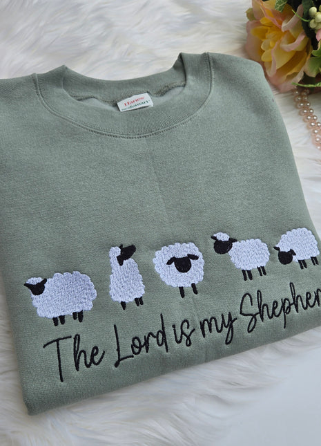 Green sweatshirt with embroidered sheep and text on a white surface with flowers and jewelry.