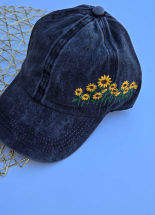 A dark washed denim baseball cap featuring embroidered yellow sunflowers with green stems on the side, displayed on a blue background next to a gold wire mesh tray.