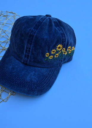 A dark blue washed cotton baseball cap with embroidered yellow flowers and green stems on the side, displayed on a solid blue background next to a gold wire mesh prop.