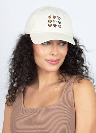 A woman with curly dark hair wearing a cream-colored baseball cap with embroidered hearts in brown, beige, and white, paired with a ribbed brown tank top against a light gray background.