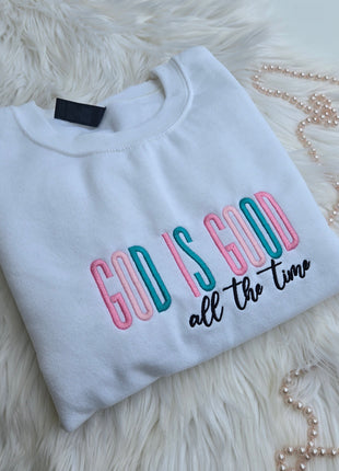 A folded white sweatshirt with colorful embroidered text reading 'God is Good All the Time '
