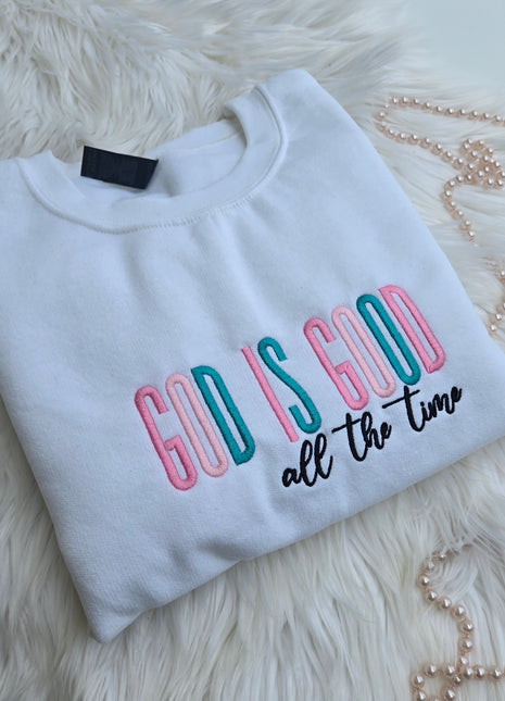A folded white sweatshirt with colorful embroidered text reading 'God is Good All the Time '