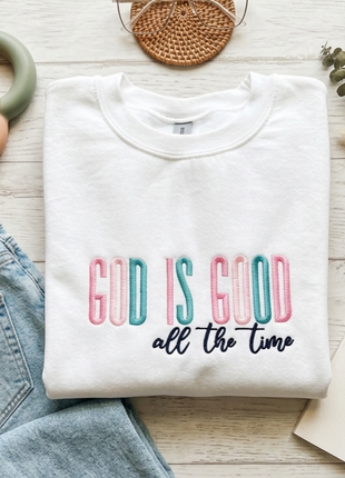 A folded white crewneck sweatshirt with the text 'GOD IS GOOD all the time' printed on the front, styled in a flat lay with blue jeans