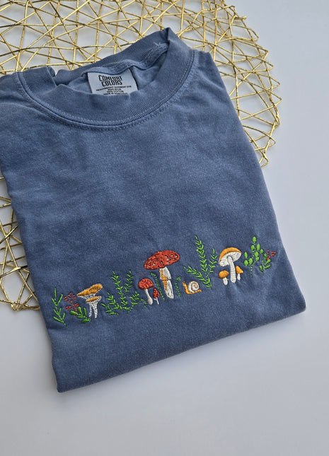 A folded slate blue t-shirt with a vertical embroidered line of colorful mushrooms and green leaves, displayed on a gold wire geometric mat against a light gray background.