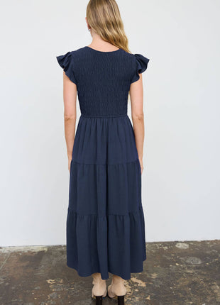 A woman viewed from behind wearing a navy blue midi dress with ruffled cap sleeves, a smocked bodice, and a tiered skirt, paired with beige high-heeled sandals against a white wall.