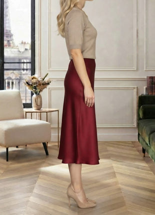 A woman stands in profile wearing a beige short-sleeve top and a satin-like burgundy midi skirt in a styled interior room with wood flooring, a chair, and a sofa.