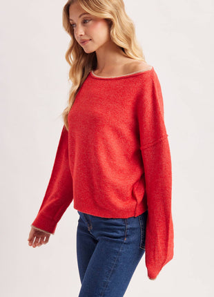 A woman wearing a bright red off-the-shoulder knit sweater and blue jeans, photographed against a clean light gray studio background.