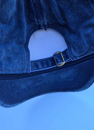Close-up of the back of a blue denim cap with an adjustable fabric strap and a small gold-tone metal buckle, set against a solid blue background.