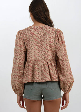 A woman shown from the back wearing a long-sleeve peplum blouse with a dense small floral print in brown and beige, paired with sage green denim shorts, against a plain white studio background.