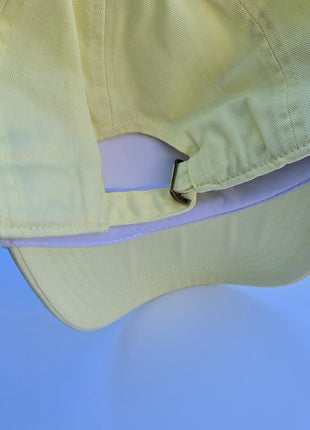 A pale yellow baseball cap shown from a close-up rear angle, highlighting the fabric texture, stitching, and adjustable snapback closure against a solid blue background.