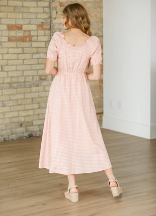 Woman wearing a pink checkered dress standing in a room with wooden flooring and a brick wall.