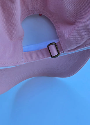 Close-up of the back of a dusty pink baseball cap, showing the fabric texture, the adjustable strap, and the black metal slider buckle against a solid blue background.
