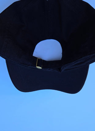 A black baseball cap with a wide-brimmed sun visor, shown from the back to display the adjustable black fabric strap with a small gold metal buckle, set against a solid blue background.